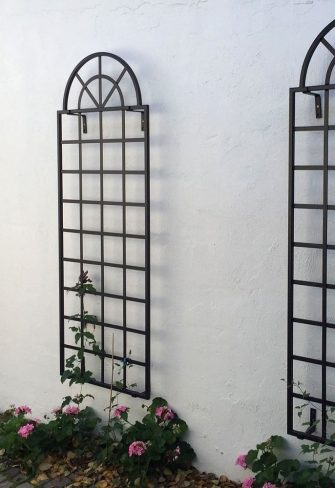 Three metal Orangery Wall Trellises by Classic Garden Elements, mounted on a white wall and planted with clematis and geraniums